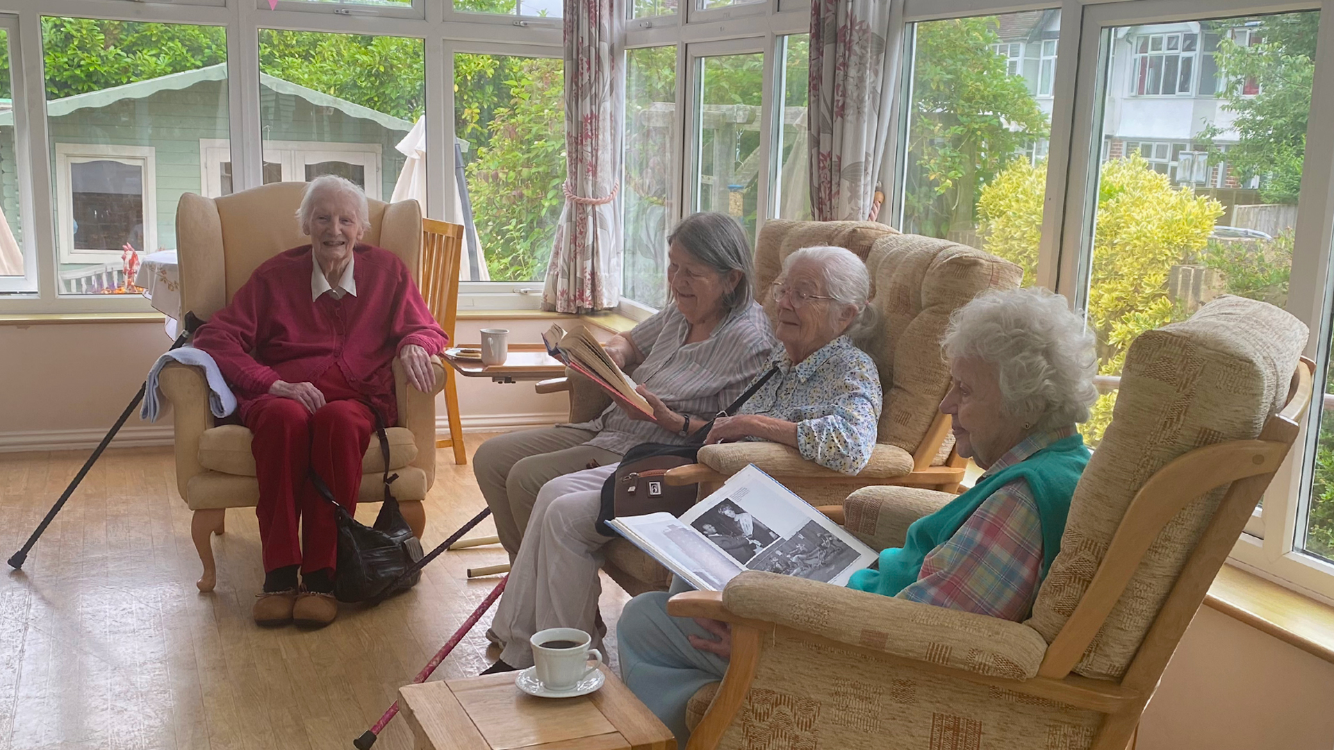 Four care home residents smile and chat in the sunny conservatory, reading books, sat with cups of tea and biscuits